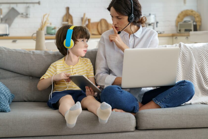 Nervous mother sitting on couch at home during lockdown, work on laptop, child distracts from work