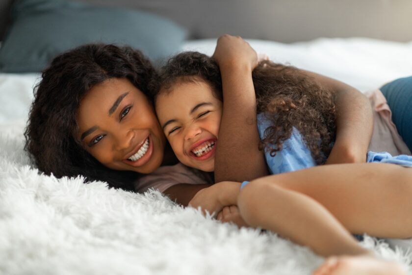 Parent child love. Happy young black woman embracing her little daughter on bed, cute girl laughing