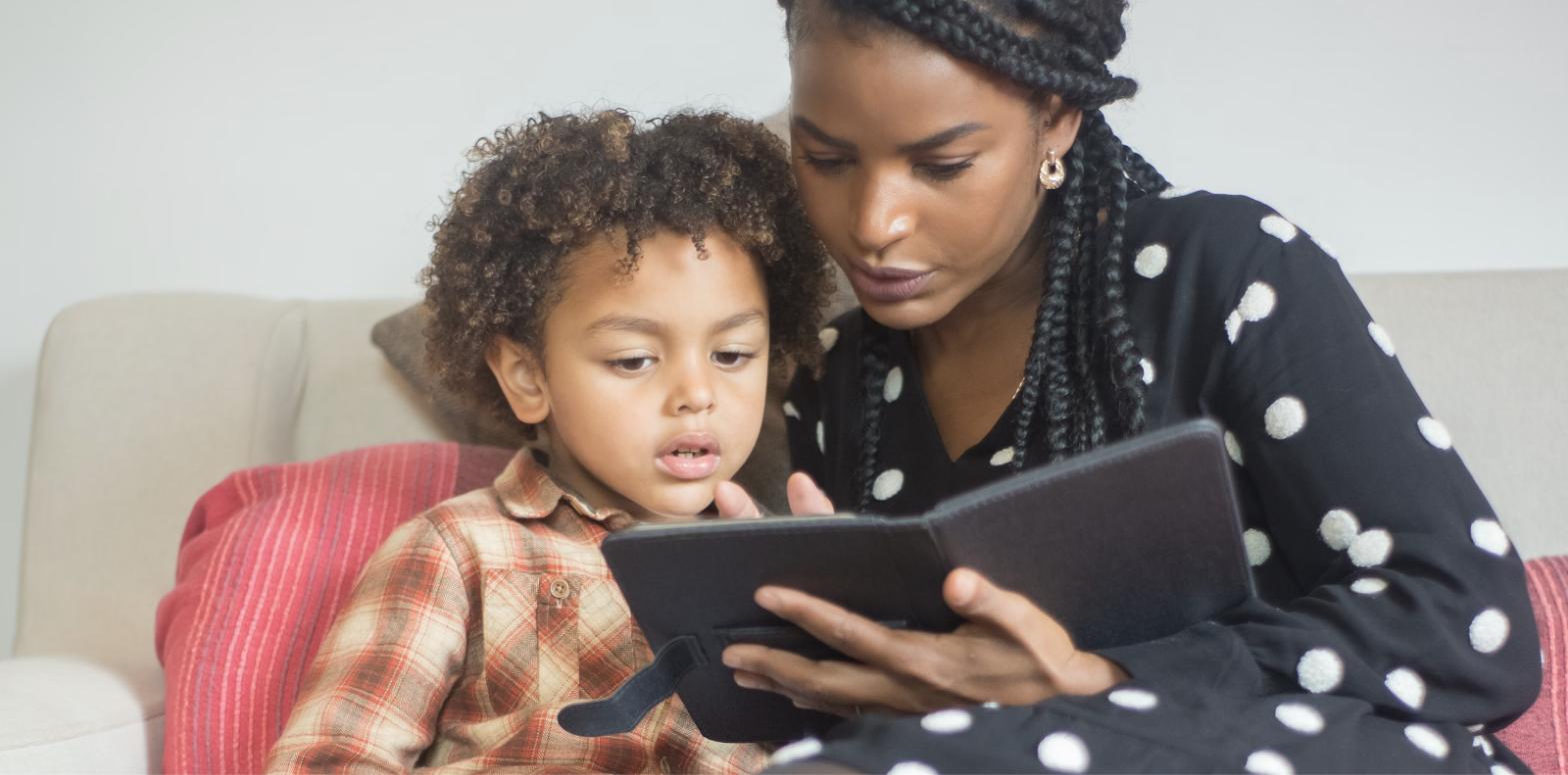 A woman and a young child sitting close together on a couch, both looking at a tablet she is holding