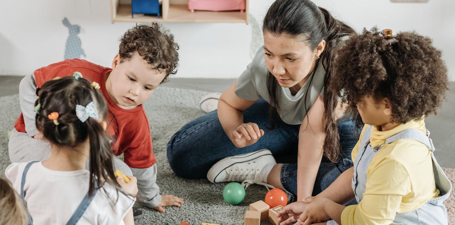 An early childhood teacher sitting on the classroom floor, leaning in as three young children play with balls and wooden blocks